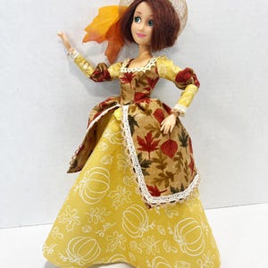May include: A doll with shoulder-length brown hair wearing a gown featuring a yellow skirt with white line drawings of pumpkins and leaves, and a brown bodice and overskirt patterned with autumn leaves. The doll wears a tan mesh hat with a leaf.