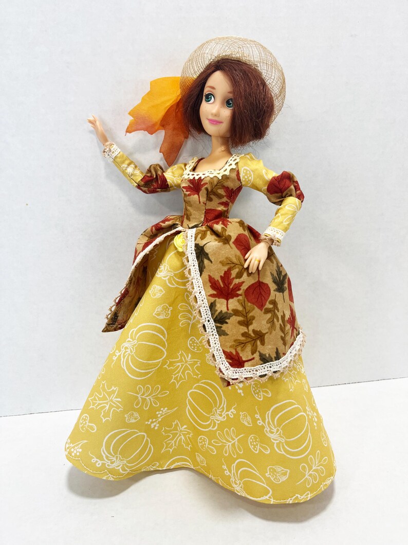 May include: A doll with shoulder-length brown hair wearing a gown featuring a yellow skirt with white line drawings of pumpkins and leaves, and a brown bodice and overskirt patterned with autumn leaves. The doll wears a tan mesh hat with a leaf.