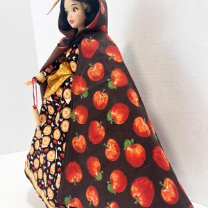 Op de afbeelding: Een pop met zwart haar draagt een bruine cape met capuchon met een rode appelprint over een zwarte jurk met een taartpatroon. De pop heeft een gele sjerp in de taille en houdt een klein houten accessoire aan een rood touwtje vast.