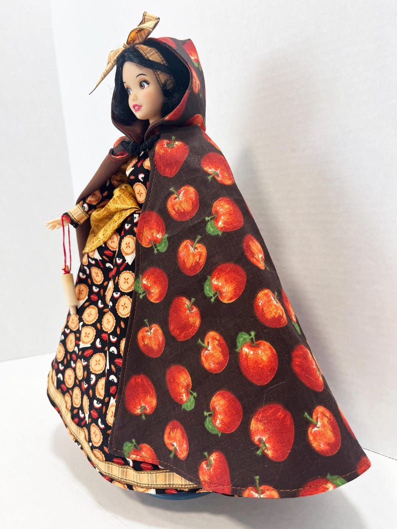 Op de afbeelding: Een pop met zwart haar draagt een bruine cape met capuchon met een rode appelprint over een zwarte jurk met een taartpatroon. De pop heeft een gele sjerp in de taille en houdt een klein houten accessoire aan een rood touwtje vast.