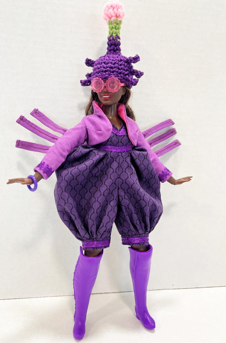 May include: A doll dressed in a purple ensemble, featuring a patterned jumpsuit, bolero jacket, and knee-high boots. The doll also wears a distinctive hat, pink glasses, and a purple bracelet. The outfit is a unique design.