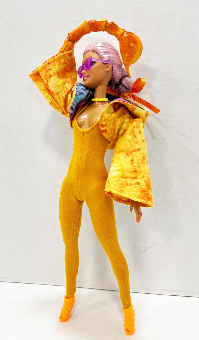 K&ouml;nnte beinhalten: Eine Puppe mit hellrosa Haar tr&auml;gt eine violette Sonnenbrille, einen gelben Body und eine gelbe Jacke mit orangefarbenem Fleckenmuster. Die Puppe hat ein gelbes Halsband und orangefarbene Schuhe sowie einen gro&szlig;en Skorpionschwanz als Accessoire.