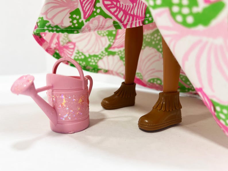 May include: A pink miniature watering can with glitter accents sits next to brown doll boots. The doll's legs are visible, and a colorful floral patterned skirt in pink, green, and white is in the background.