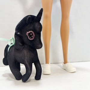 May include: A black flocked unicorn figurine with a single horn, a light green ribbon tied around its neck, and a large eye featuring a pink iris with a star detail. The figurine stands next to a pair of light-skinned legs wearing white shoes.