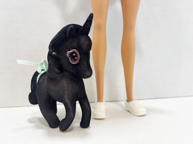 May include: A black flocked unicorn figurine with a single horn, a light green ribbon tied around its neck, and a large eye featuring a pink iris with a star detail. The figurine stands next to a pair of light-skinned legs wearing white shoes.