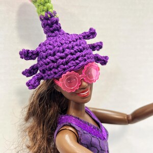 May include: A doll wearing a purple crocheted hat with a pink and green top, and pink flower-shaped sunglasses. The doll is also wearing a purple dress. The hat has multiple protrusions and a bulbous top.