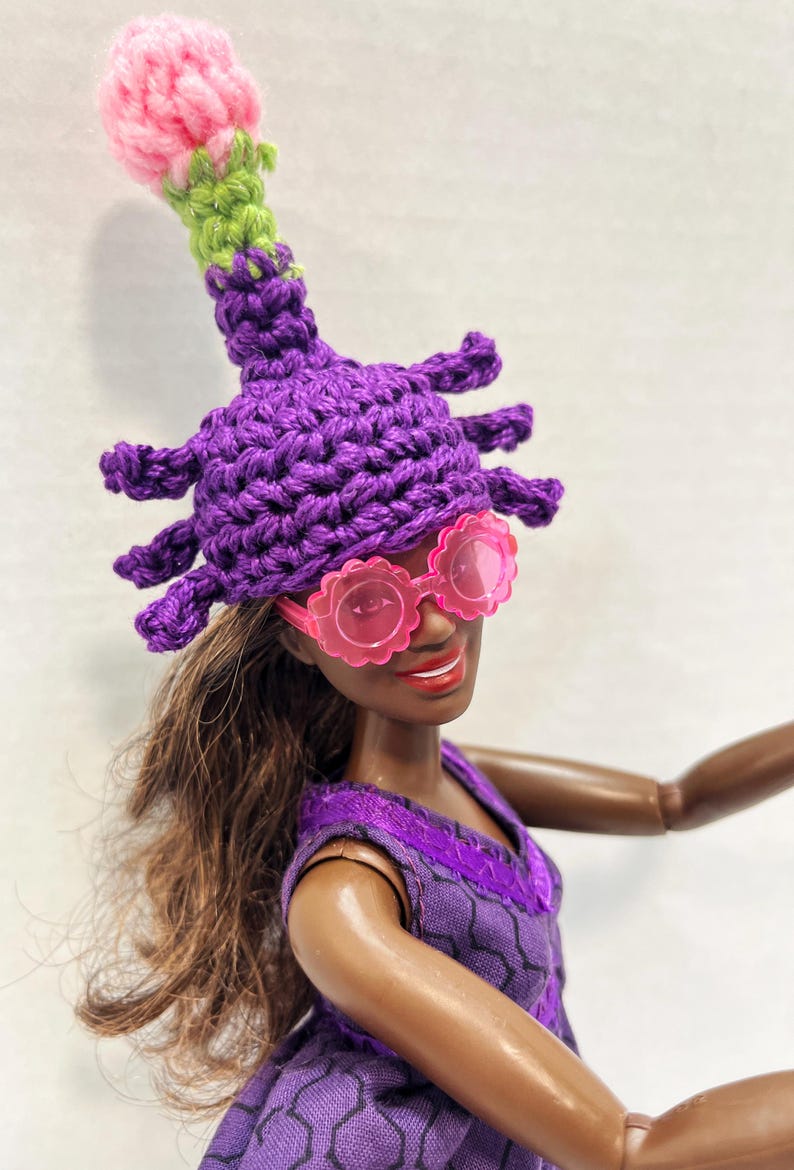 May include: A doll wearing a purple crocheted hat with a pink and green top, and pink flower-shaped sunglasses. The doll is also wearing a purple dress. The hat has multiple protrusions and a bulbous top.