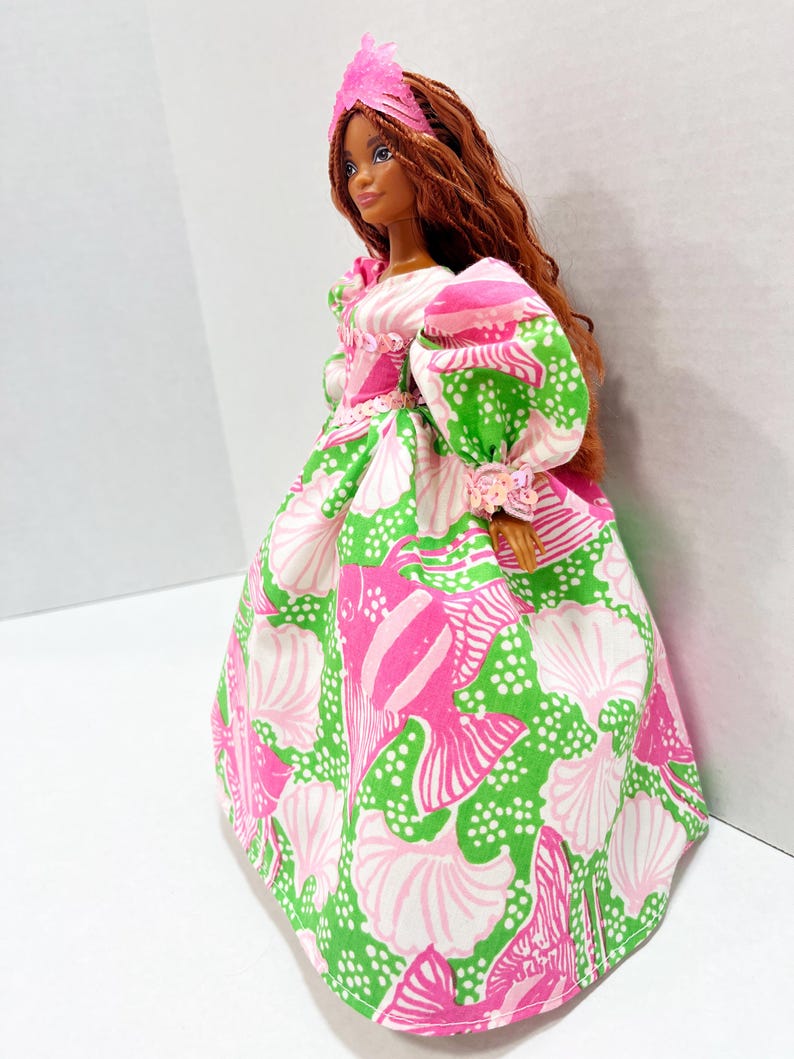 May include: A fashion doll wearing a vibrant, patterned gown. The dress features a pink, green, and white seashell design. The doll has long, auburn hair and a pink headpiece. The sleeves are puffed, and the dress has a full skirt.
