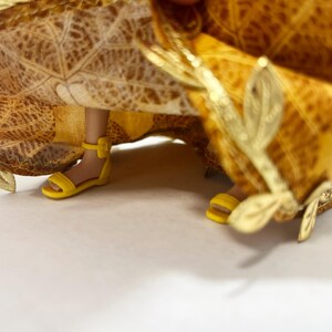 May include: A doll wearing a yellow and brown leaf-patterned skirt with gold leaf-shaped embellishments along the hem. The doll has light brown skin and is wearing bright yellow strappy sandals with an ankle strap and a small buckle.