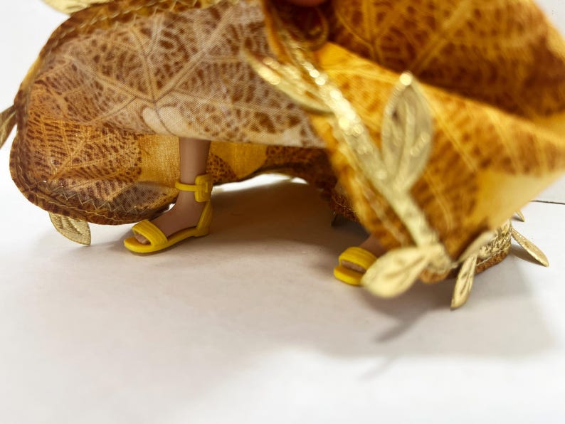 May include: A doll wearing a yellow and brown leaf-patterned skirt with gold leaf-shaped embellishments along the hem. The doll has light brown skin and is wearing bright yellow strappy sandals with an ankle strap and a small buckle.
