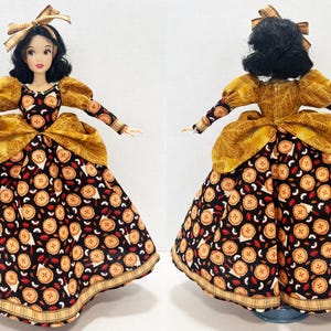 Op de afbeelding: Een pop met zwart haar gestyled met een geruite strik, gekleed in een jurk met lange mouwen van zwarte stof met een herhalend patroon van kleine taartjes en rode lippen. De jurk heeft een peplum van goudkleurige, textuurrijke stof en een geruite zoom.