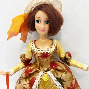 May include: A doll with shoulder-length auburn hair wearing a tan hat with an orange leaf accent. The doll wears a dress featuring a pattern of red and brown autumn leaves on a tan background, accented with white lace trim and a small yellow rose.
