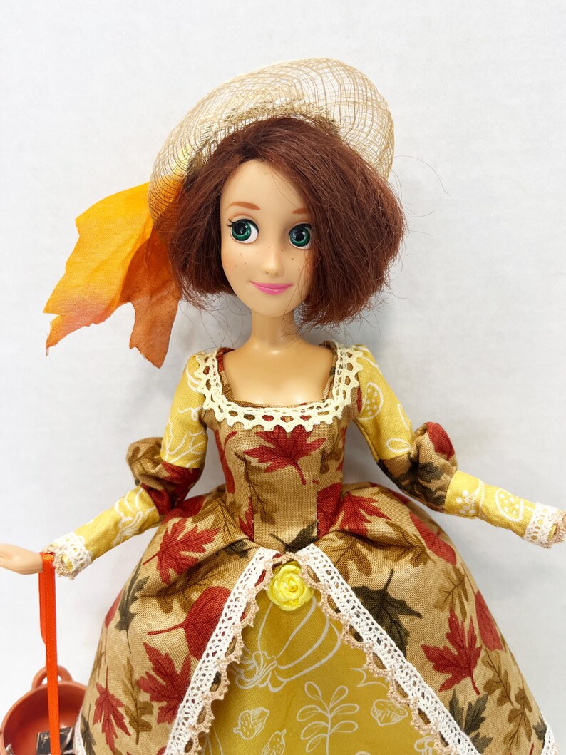 May include: A doll with shoulder-length auburn hair wearing a tan hat with an orange leaf accent. The doll wears a dress featuring a pattern of red and brown autumn leaves on a tan background, accented with white lace trim and a small yellow rose.