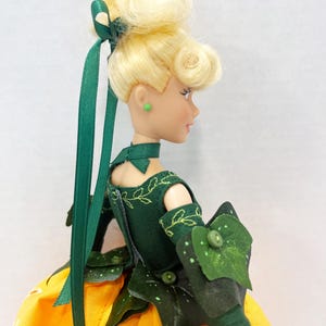 May include: A doll with blonde hair styled in an updo with a green ribbon, wearing a dark green bodice with embroidered leaf patterns, a choker, and green arm sleeves with leaf accents. The doll has a yellow skirt with white floral patterns.