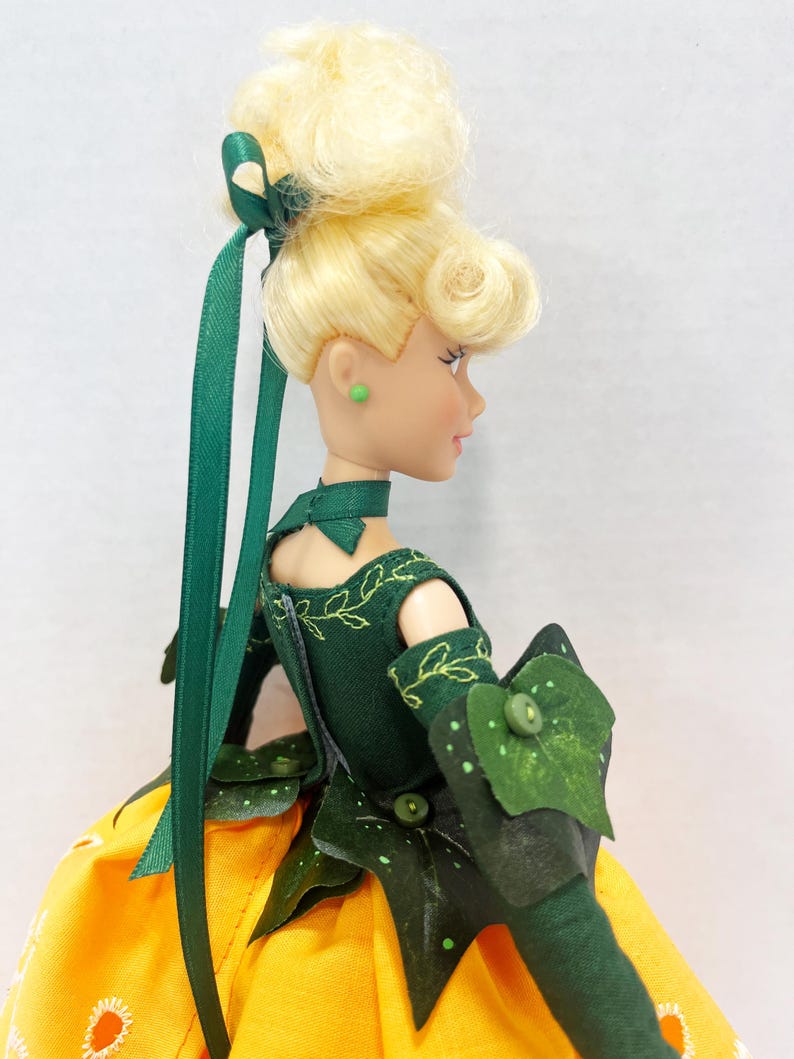 May include: A doll with blonde hair styled in an updo with a green ribbon, wearing a dark green bodice with embroidered leaf patterns, a choker, and green arm sleeves with leaf accents. The doll has a yellow skirt with white floral patterns.