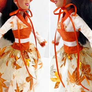 May include: A doll wearing a white long-sleeved top with orange leaf patterns, an orange bodice, and a white skirt featuring metallic orange leaf appliques. The doll has long black hair and wears an orange hat tied with orange ribbons under the chin.