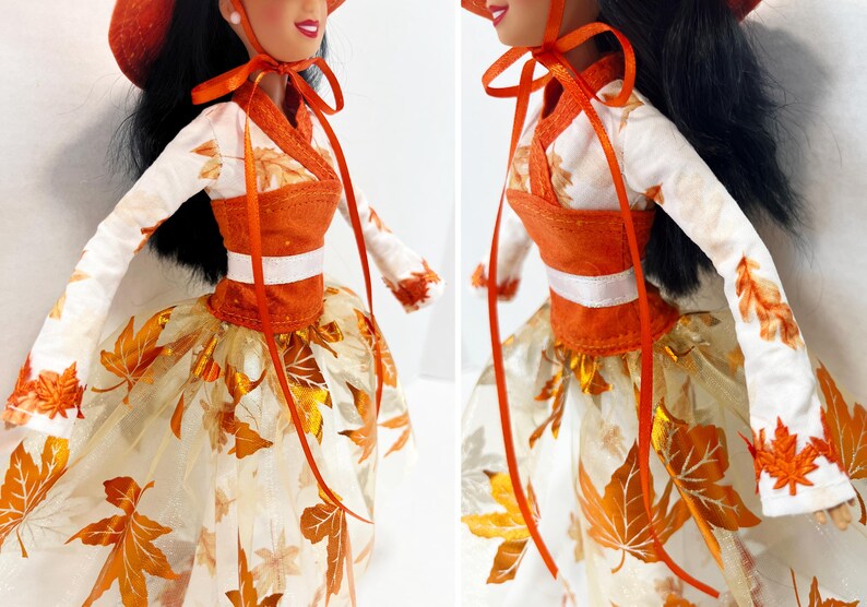 May include: A doll wearing a white long-sleeved top with orange leaf patterns, an orange bodice, and a white skirt featuring metallic orange leaf appliques. The doll has long black hair and wears an orange hat tied with orange ribbons under the chin.