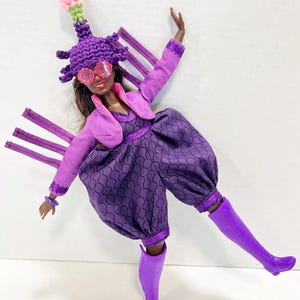 May include: A doll in a purple ensemble, featuring a patterned jumpsuit, bolero jacket, and distinctive hat. The doll wears pink glasses and purple boots. The hat has a pink and green accent. The doll's outfit is completed with a purple bracelet.