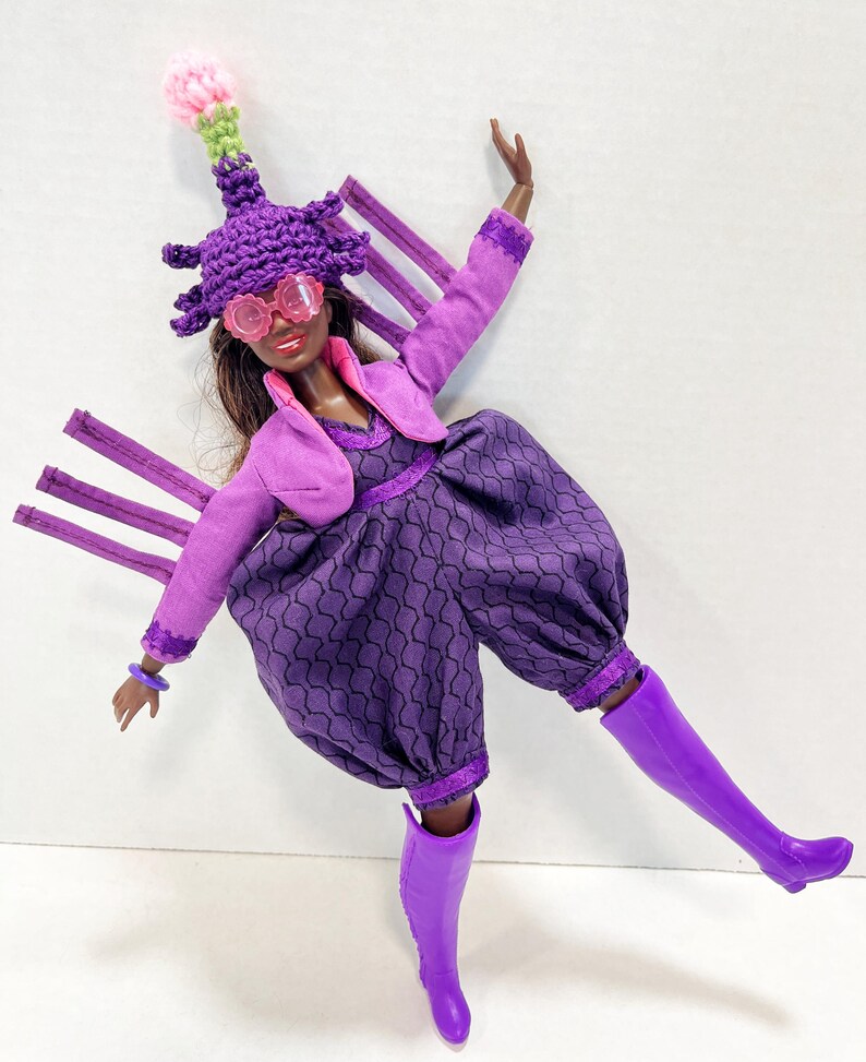 May include: A doll in a purple ensemble, featuring a patterned jumpsuit, bolero jacket, and distinctive hat. The doll wears pink glasses and purple boots. The hat has a pink and green accent. The doll's outfit is completed with a purple bracelet.