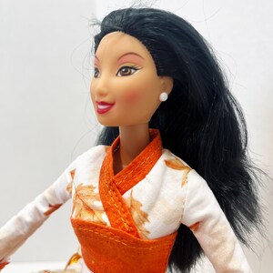 May include: A doll with long black hair, brown eyes, and red lipstick, wearing a white top featuring an orange leaf pattern with a solid orange wrap-style bodice and a white waistband. The doll has small white stud earrings and a fair complexion.