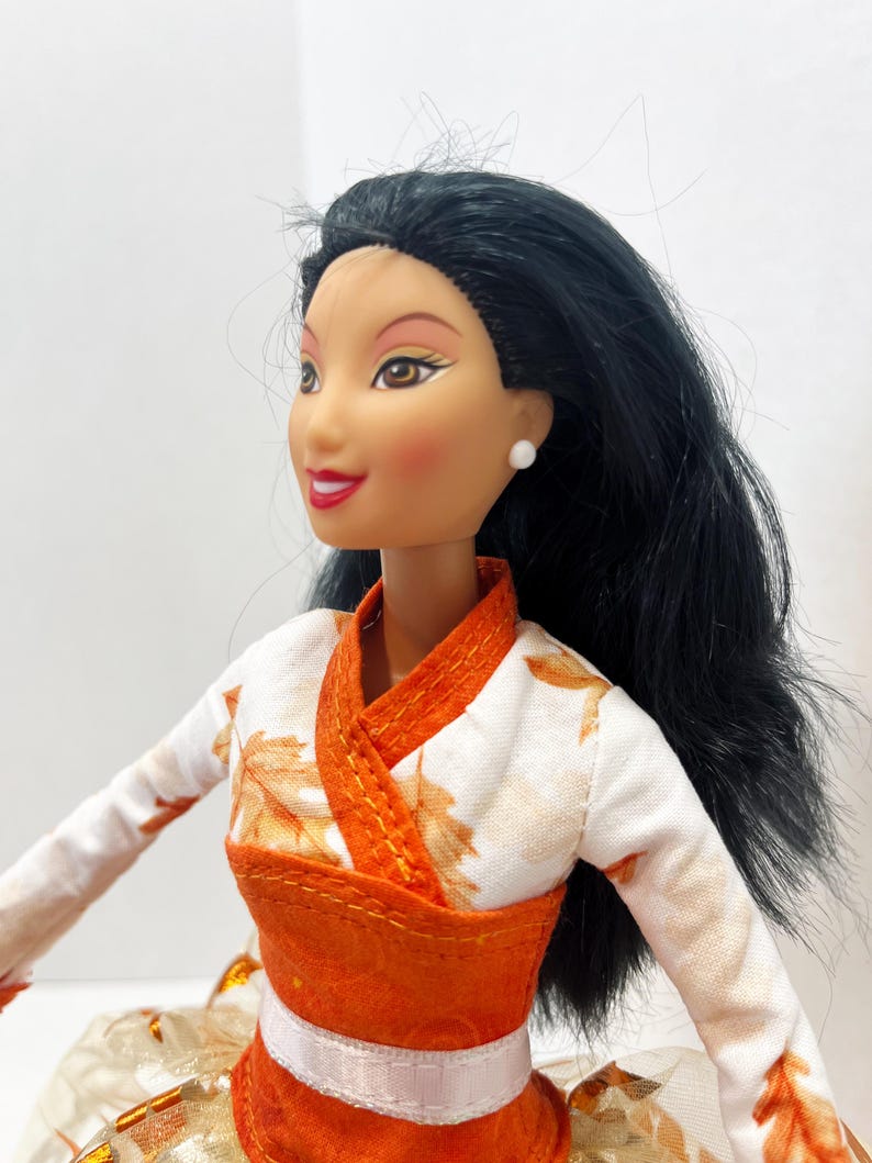 May include: A doll with long black hair, brown eyes, and red lipstick, wearing a white top featuring an orange leaf pattern with a solid orange wrap-style bodice and a white waistband. The doll has small white stud earrings and a fair complexion.