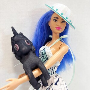 May include: A doll with long blue hair wearing a white hat with teal hearts and a white dress featuring a grid pattern and polyhedral dice graphics. The doll holds a small, black, textured creature figure with large pink eyes and a single horn.