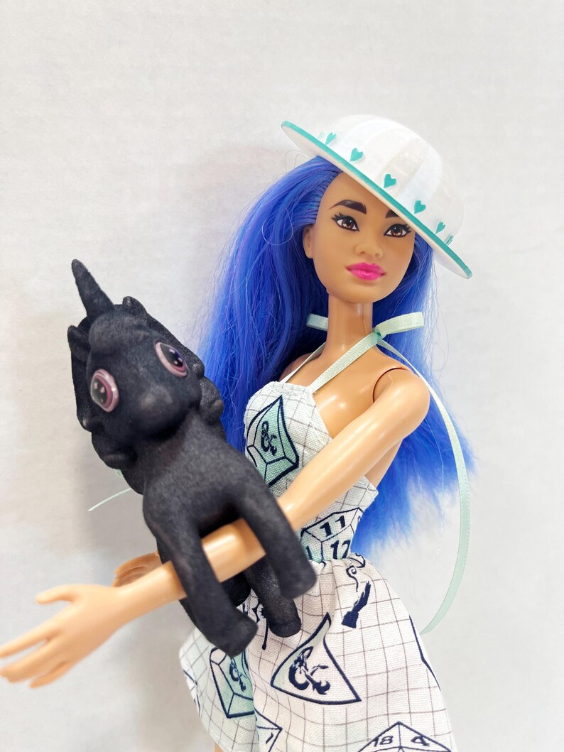 May include: A doll with long blue hair wearing a white hat with teal hearts and a white dress featuring a grid pattern and polyhedral dice graphics. The doll holds a small, black, textured creature figure with large pink eyes and a single horn.