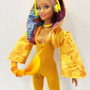 K&ouml;nnte beinhalten: Eine Puppe mit hellviolettem Haar tr&auml;gt einen gelben Body und eine gelbe Kapuzenjacke mit orange-gelbem Batikmuster. Das Innenfutter der Kapuze hat ein blau-violettes Muster. Die Puppe tr&auml;gt einen gelben Kopfh&ouml;rer und ein gelbes Halsband.