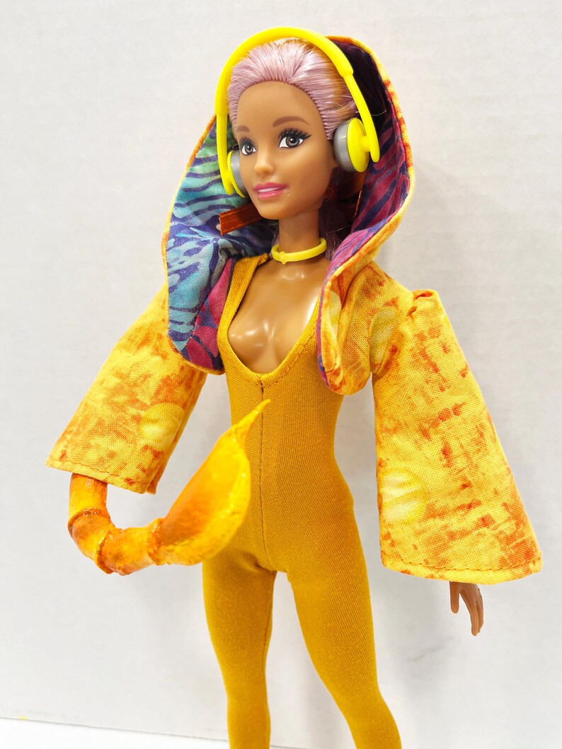 K&ouml;nnte beinhalten: Eine Puppe mit hellviolettem Haar tr&auml;gt einen gelben Body und eine gelbe Kapuzenjacke mit orange-gelbem Batikmuster. Das Innenfutter der Kapuze hat ein blau-violettes Muster. Die Puppe tr&auml;gt einen gelben Kopfh&ouml;rer und ein gelbes Halsband.
