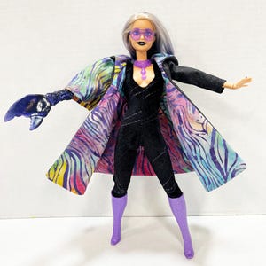 May include: A fashion doll in a black jumpsuit, purple boots, and sunglasses. The doll wears a colorful coat with a zebra stripe pattern and a purple necklace. The doll has silver hair with blue highlights and a dark lip color.