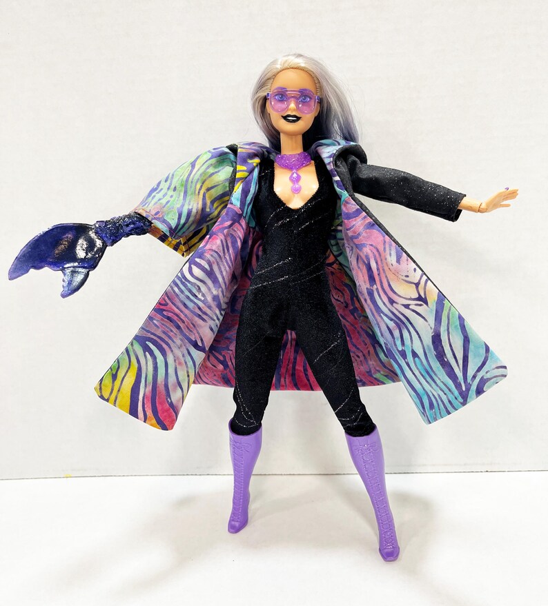 May include: A fashion doll in a black jumpsuit, purple boots, and sunglasses. The doll wears a colorful coat with a zebra stripe pattern and a purple necklace. The doll has silver hair with blue highlights and a dark lip color.