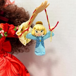 May include: A small, hand-painted doll ornament featuring a blonde-haired character in a blue dress with white polka dots. The doll is suspended by a red and gold string, with a gold crown. The doll is held by a doll hand.