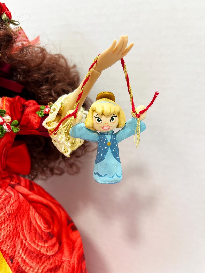 May include: A small, hand-painted doll ornament featuring a blonde-haired character in a blue dress with white polka dots. The doll is suspended by a red and gold string, with a gold crown. The doll is held by a doll hand.
