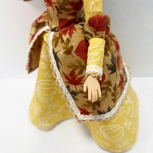 May include: A doll wearing a dress with a brown bodice featuring a red and brown autumn leaf print, paired with a yellow skirt decorated with white pumpkin outlines. The dress includes cream-colored lace trim at the neckline and cuffs.