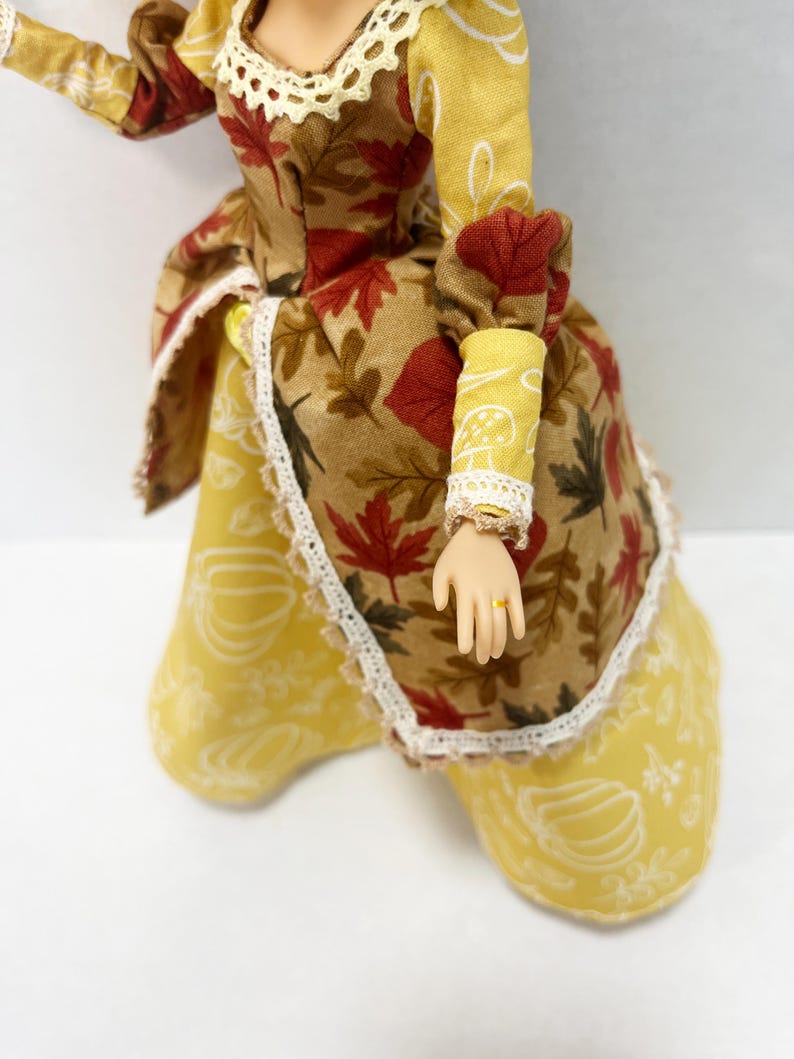 May include: A doll wearing a dress with a brown bodice featuring a red and brown autumn leaf print, paired with a yellow skirt decorated with white pumpkin outlines. The dress includes cream-colored lace trim at the neckline and cuffs.