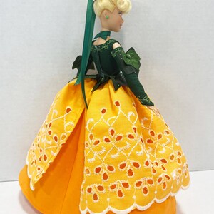 May include: A doll with blonde hair styled in an updo with a green ribbon. The doll wears a dark green long-sleeved bodice with leaf-like accents and a bright orange skirt featuring a white eyelet lace overlay with a scalloped hemline.