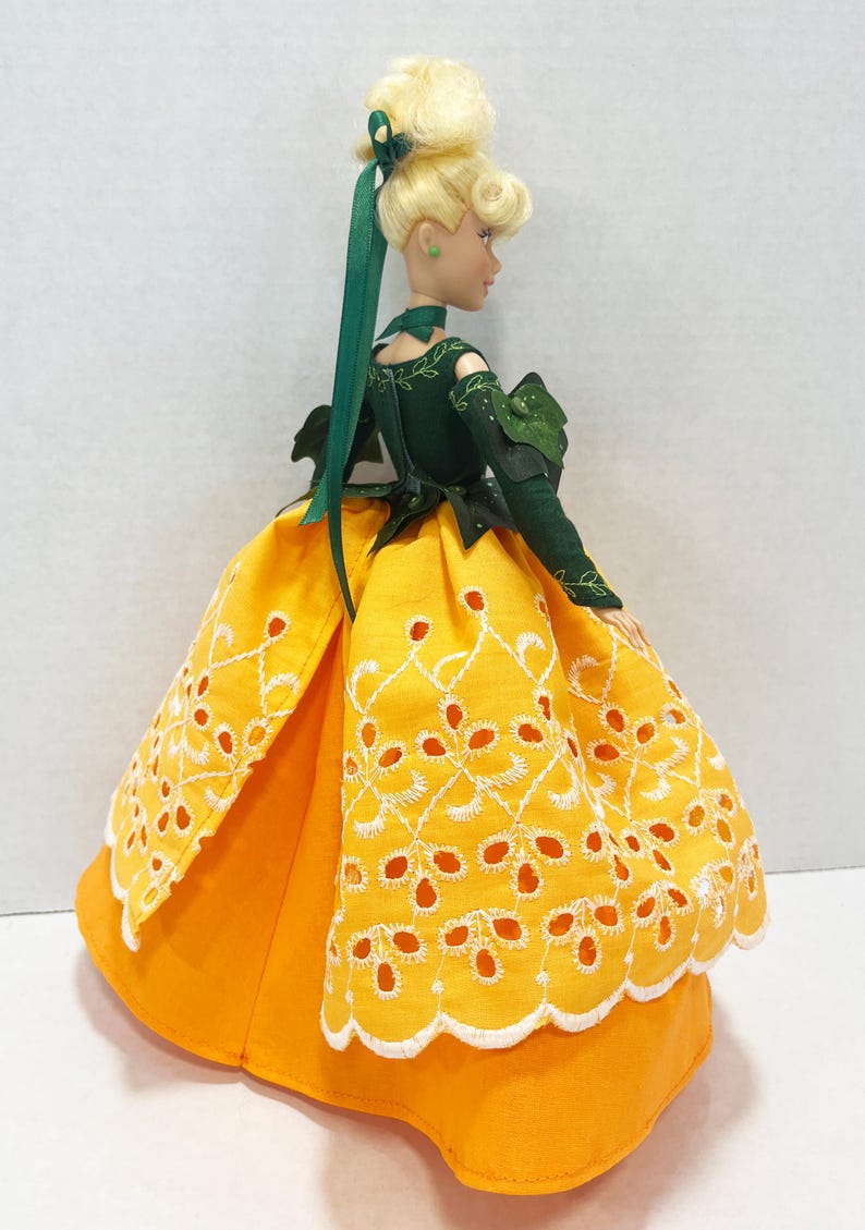 May include: A doll with blonde hair styled in an updo with a green ribbon. The doll wears a dark green long-sleeved bodice with leaf-like accents and a bright orange skirt featuring a white eyelet lace overlay with a scalloped hemline.