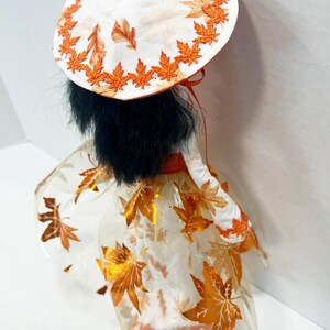 May include: A doll with black hair wearing a white hat and dress featuring an orange maple leaf pattern. The hat has an orange embroidered leaf border, and the dress includes a sheer overlay with metallic orange leaf accents and a solid orange waist.