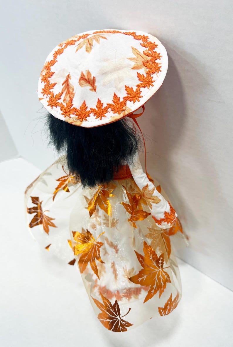 May include: A doll with black hair wearing a white hat and dress featuring an orange maple leaf pattern. The hat has an orange embroidered leaf border, and the dress includes a sheer overlay with metallic orange leaf accents and a solid orange waist.