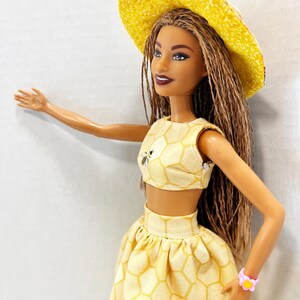 Puede incluir: Mu&ntilde;eca con un atuendo amarillo con un patr&oacute;n de panal y gr&aacute;ficos de abejas. El atuendo incluye un top corto, una falda y un sombrero de ala ancha. Una pulsera de flores rosas adorna la mu&ntilde;eca. La mu&ntilde;eca tiene pelo largo casta&ntilde;o.