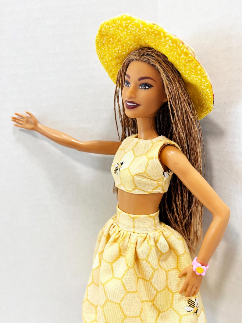 Puede incluir: Mu&ntilde;eca con un atuendo amarillo con un patr&oacute;n de panal y gr&aacute;ficos de abejas. El atuendo incluye un top corto, una falda y un sombrero de ala ancha. Una pulsera de flores rosas adorna la mu&ntilde;eca. La mu&ntilde;eca tiene pelo largo casta&ntilde;o.