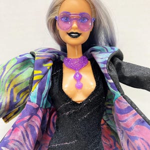 May include: A doll wearing a black dress, purple sunglasses, and a matching necklace. The doll has silver and blue hair and is wearing a jacket with a colorful, patterned lining. The jacket is black on the outside.