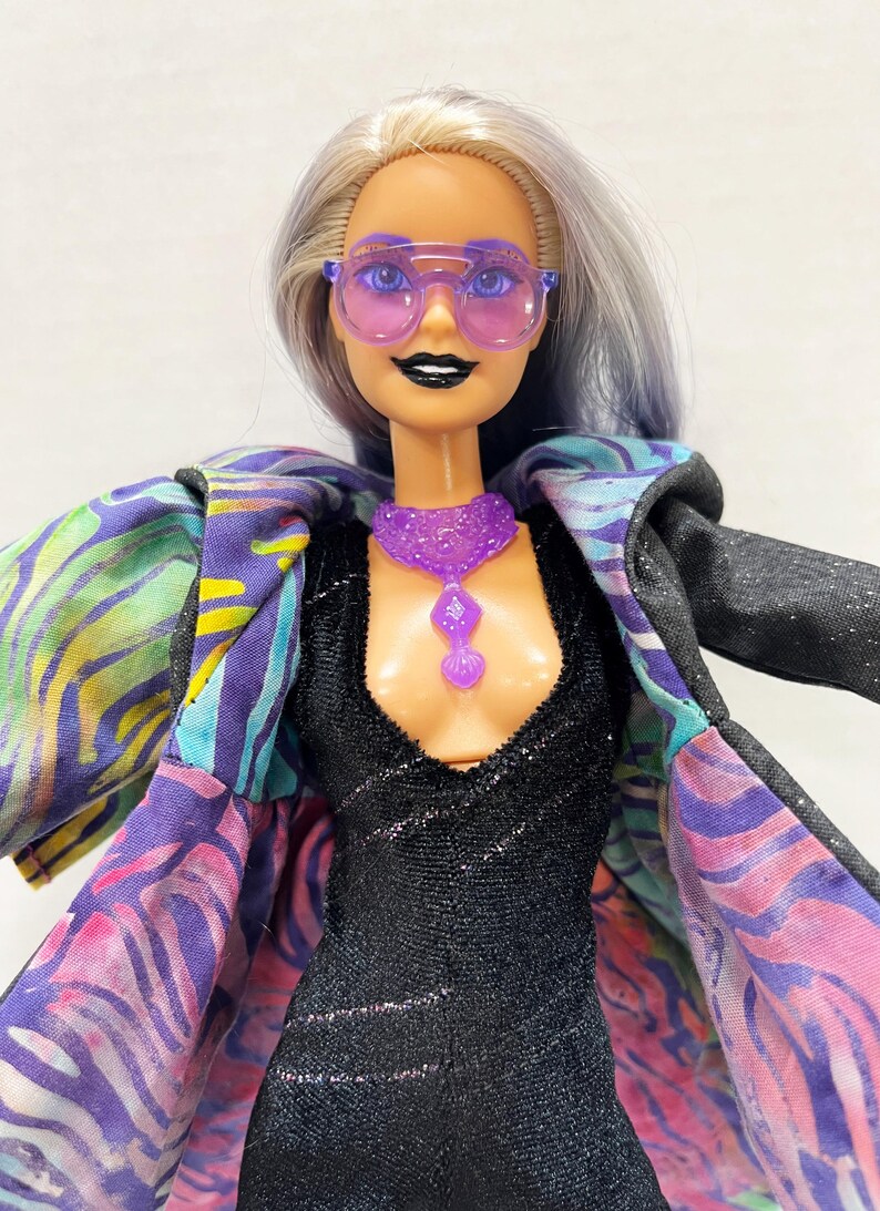 May include: A doll wearing a black dress, purple sunglasses, and a matching necklace. The doll has silver and blue hair and is wearing a jacket with a colorful, patterned lining. The jacket is black on the outside.