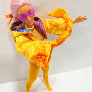 K&ouml;nnte beinhalten: Eine Puppe mit rosa Haaren, die eine durchscheinende violette Sonnenbrille, ein gelbes Halsband und eine gelbe Kapuzenjacke mit mehrfarbigem Innenfutter tr&auml;gt. Die Puppe hat einen gro&szlig;en, gl&auml;nzenden orangefarbenen Skorpion-Greifarm an einem Arm.