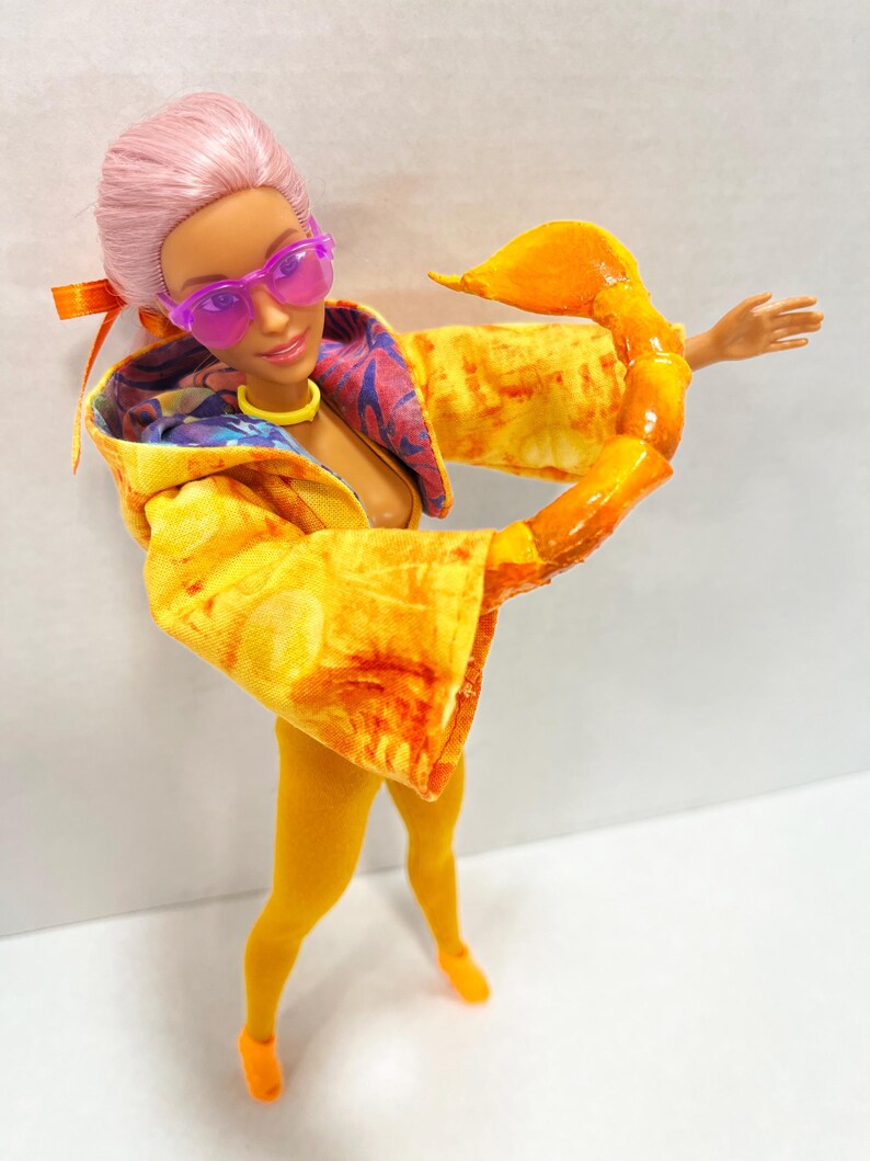 K&ouml;nnte beinhalten: Eine Puppe mit rosa Haaren, die eine durchscheinende violette Sonnenbrille, ein gelbes Halsband und eine gelbe Kapuzenjacke mit mehrfarbigem Innenfutter tr&auml;gt. Die Puppe hat einen gro&szlig;en, gl&auml;nzenden orangefarbenen Skorpion-Greifarm an einem Arm.