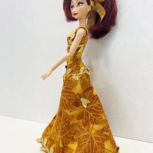May include: A doll with dark red hair styled in a high ponytail with a gold ribbon. The doll wears a floor-length sleeveless dress featuring a brown and yellow maple leaf pattern, accented with metallic gold leaf trim at the waist and hem.
