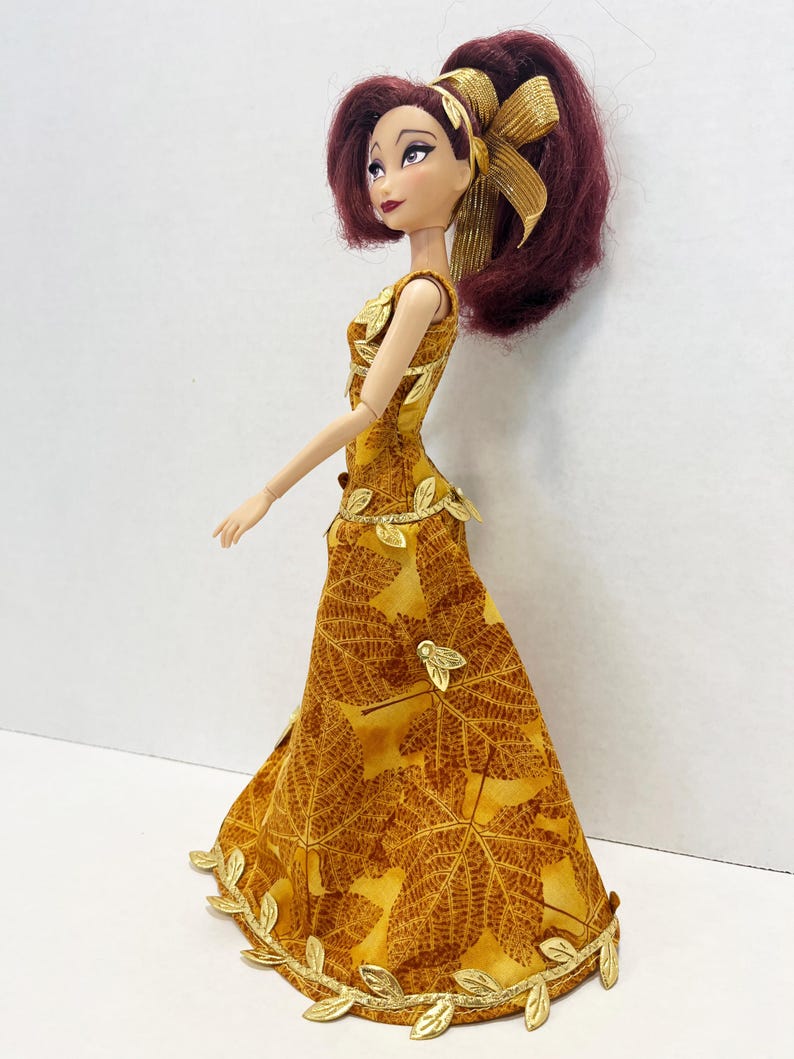 May include: A doll with dark red hair styled in a high ponytail with a gold ribbon. The doll wears a floor-length sleeveless dress featuring a brown and yellow maple leaf pattern, accented with metallic gold leaf trim at the waist and hem.