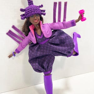 May include: A doll dressed in a purple ensemble, including a hat and boots, holding a pink dumbbell. The outfit features a patterned jumpsuit, a jacket, and a distinctive hat with a pink and green detail.