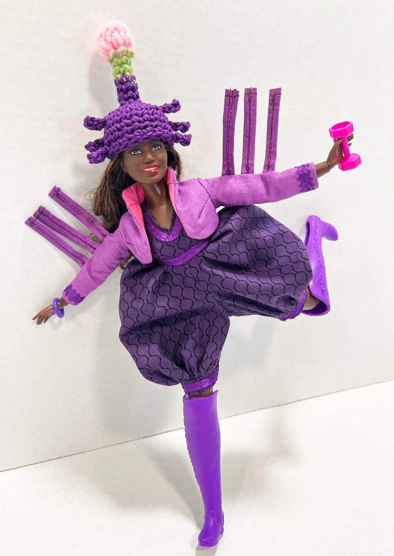 May include: A doll dressed in a purple ensemble, including a hat and boots, holding a pink dumbbell. The outfit features a patterned jumpsuit, a jacket, and a distinctive hat with a pink and green detail.