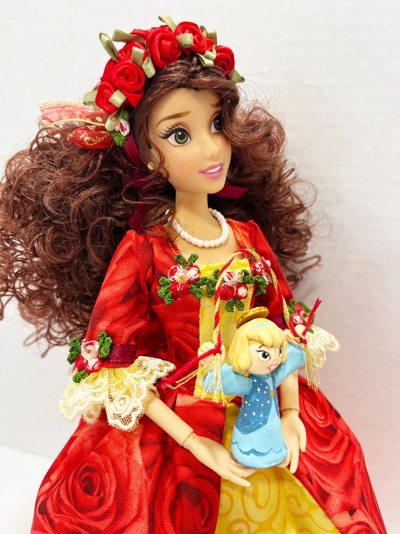 May include: A doll with brown curly hair, green eyes, and a red rose crown. The doll wears a red dress with a rose pattern, lace trim, and holds a small blue angel figurine. A pearl necklace adorns the doll's neck.